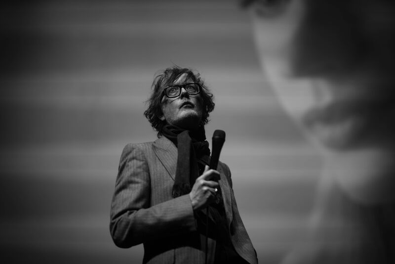 Jarvis Cocker holds a microphone while looking at the crowd in a black and white image