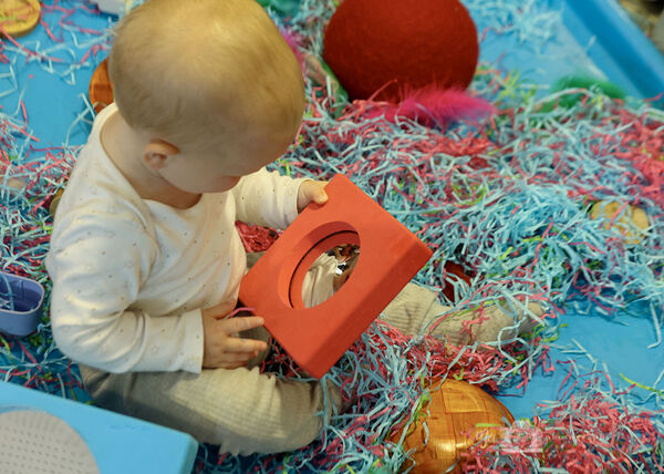 Baby looking at itself in a mirror while surrounded by shredded paper and toys