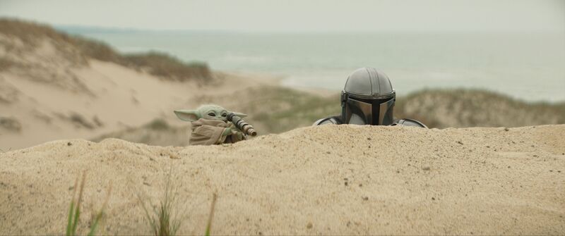 The Mandalorian and Grogu are crouched behind a sand dune. Grogu is on the left and is looking through a small telescope.