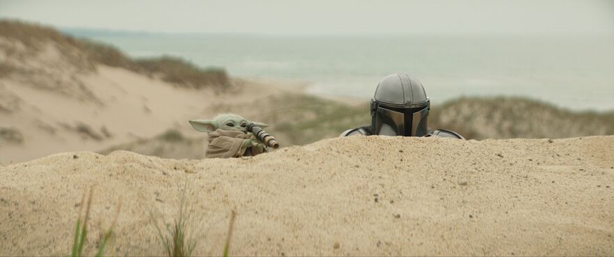The Mandalorian and Grogu are crouched behind a sand dune. Grogu is on the left and is looking through a small telescope.