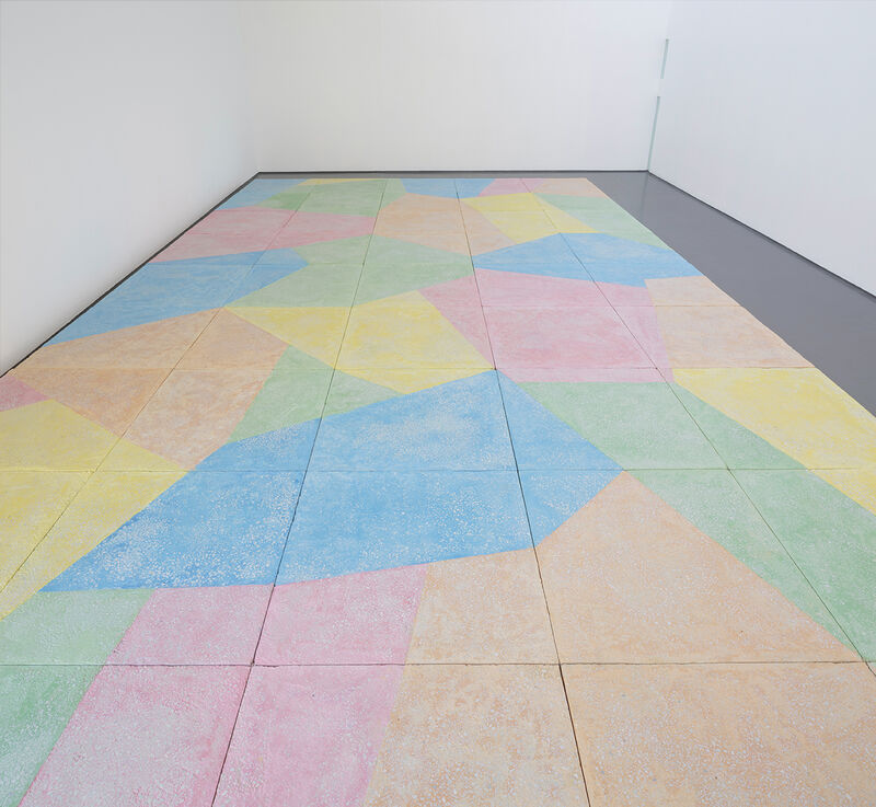 A floor based work is shown in gallery one. The walls are white and the gallery floor is painted grey. On top of this, a sculptural floor is lain in blocks, raising the floor over most of the gallery. This work is brightly coloured, with pastel blue green, yellow orange and pink, in abstract shapes.