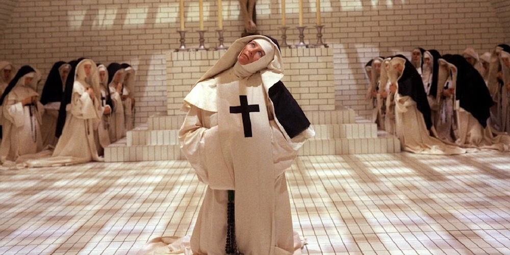 A nun kneeling on a tiled floor with other nuns praying in the background