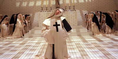 A nun kneeling on a tiled floor with other nuns praying in the background