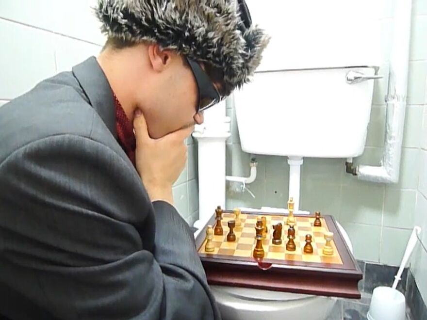 A man wearing a fluffy hat contemplates a chessboard resting on a toilet