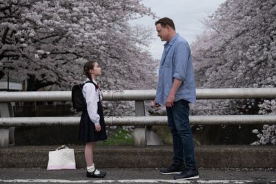 A man and a little girl stand on a bridge looking at one another, while cherry blossom blooms in the background