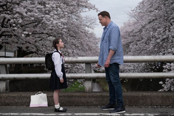 A man and a little girl stand on a bridge looking at one another, while cherry blossom blooms in the background
