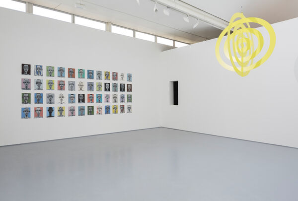 Image shows a hanging yellow artwork to the right, which is strung from the ceiling and consists of flat concentric rings. To the left we can see many small works of art ranging in bright colours which are hung in a grid on the white wall. To the corner of the gallery, in the centre of the image, is a small dark window allowing a view to the adjacent gallery. 