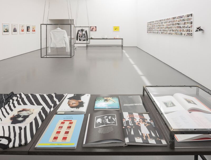 Exhibition 'Spring / Summer 2015' in the foreground, we can see a table with various 1980s fashion and music magazines open.  There is also a white jacket hanging in a. glass box, and various photographs on the wall.