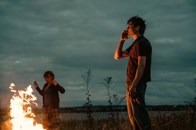 A man stands outside at dusk in front of a fire, while a woman dances next to him