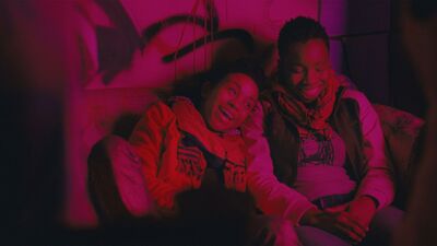 Two teenagers laugh together in a room lit with low pink lighting.