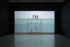 A film is shown projected in a dark space. The film still we can see shows 3 people up some scaffolding, behind them is the sea and a strip of sky that aligns perfectly with the platform. This gives the illusion of the people standing on the sea, their feet hit the horizon line.