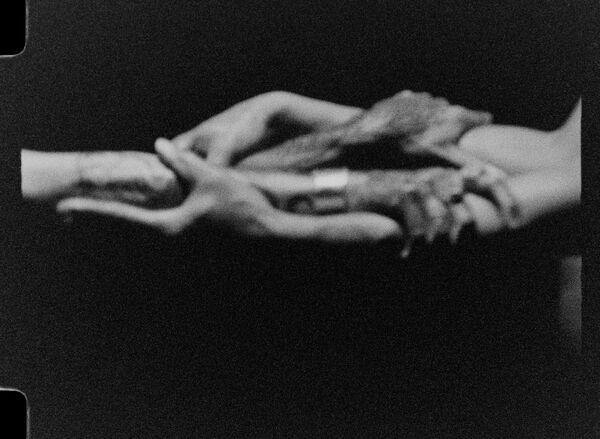A film photograph image with a black background shows interlinked hands and arms, holding each other, as if knotted together. The image is slightly blurred. 