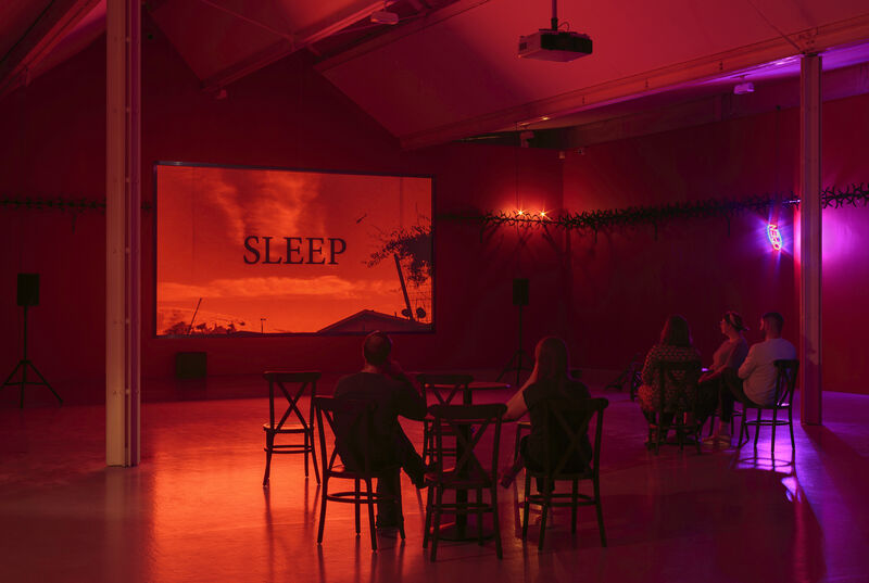 A wide shot photograph of gallery two, showing the gallery with red walls and cabaret-style seating with small tables in the floorspace. A few people sit and face a screen with a projected film. The film still is very red and shows in capital letters in a black serif font the word SLEEP. There is a bright purple light to the right of the image. 