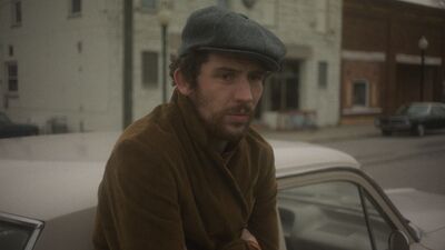 A man wearing a flatcap leans against a car on a quiet street,.