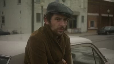 A man wearing a flatcap leans against a car on a quiet street,.