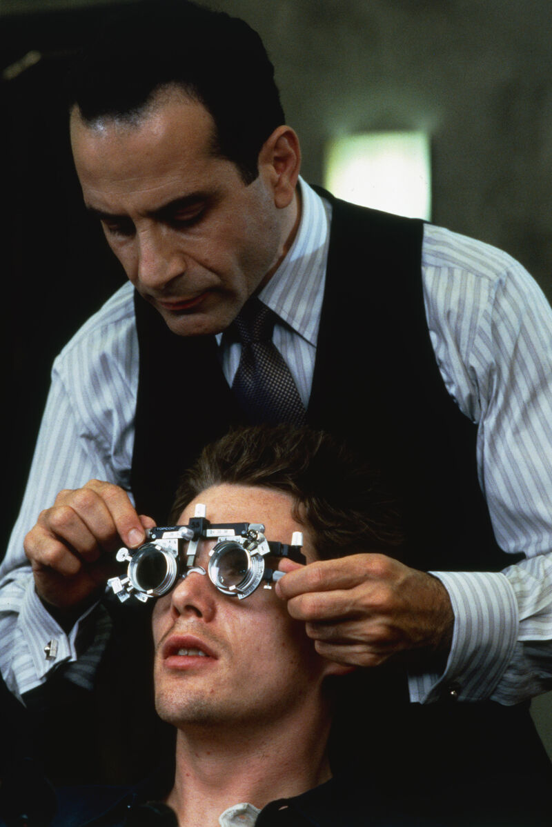 A man in a waistcoat, tie and striped shirt places metallic goggles over another man's eyes