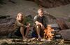 A middle-aged man and woman sit on a beach with a small fire burning.