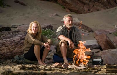 A middle-aged man and woman sit on a beach with a small fire burning.