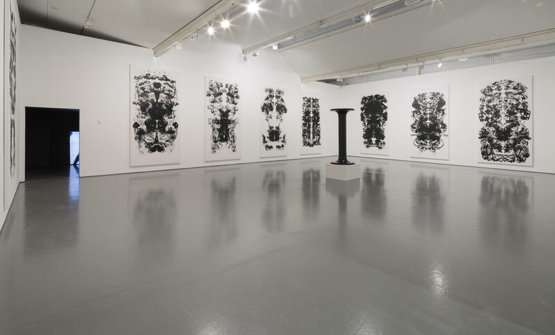 The photograph shows 8 large drawings akin to Rorschach prints, on two white walls. There is also a sculpture on a white plinth, of the letter 'I' in all black. A small opening to the left of the image goes into another room. 