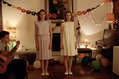 Two girls in white dresses stand looking forlornly at the camera in what a appears to be a home party scene