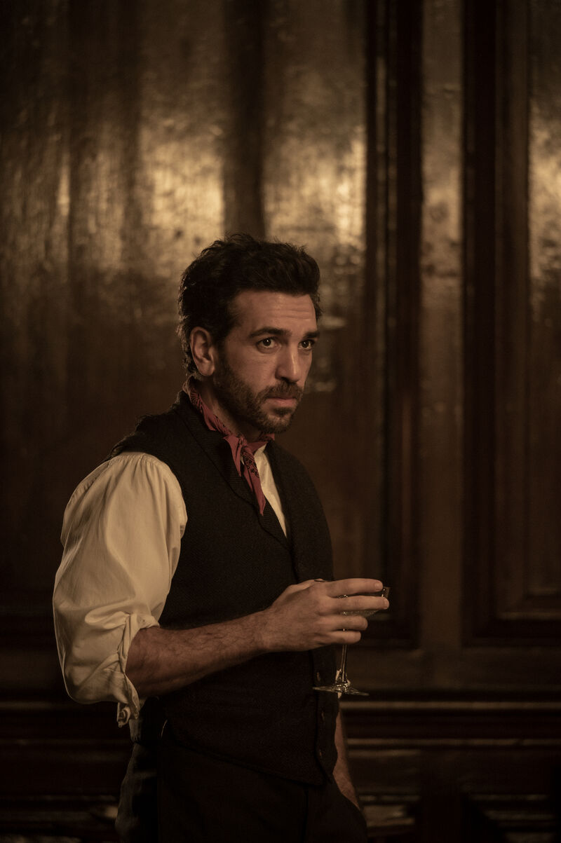 A person in period clothing stands in a dimly lit wood-panelled room, holding a small glass, with warm light reflecting across the dark walls behind them.