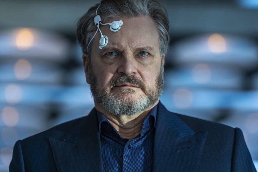 A close view of a man with electrodes stuck to his head, he has a pained expression. Colin firth is the man.