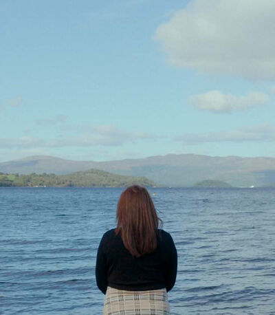 A woman with long dark hair looks at a loch