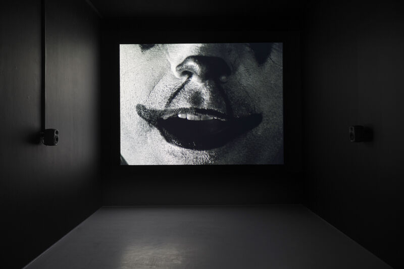 A small dark room has a moving image artwork in it. The film still shows a black and white image of a person's mouth and nose. They have makeup like a clown, with a white face and exaggerated smile. 