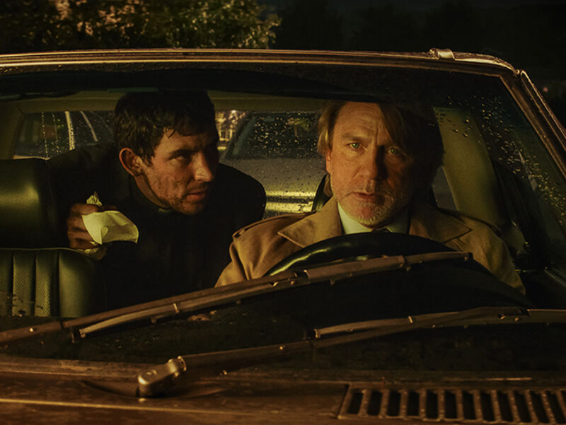 A worried man drives a vintage car while another man in the back seat leans over watching him