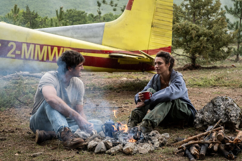 A young man and a young woman sit next to a fire in front of a propellor plane
