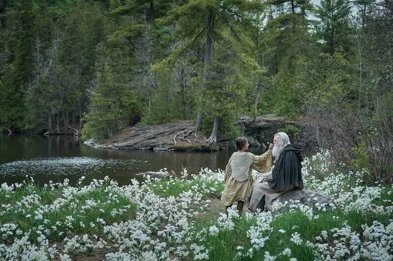An elderly man with a beard and a young woman in a dress sit next to a lake in a field surrounded by white flowers