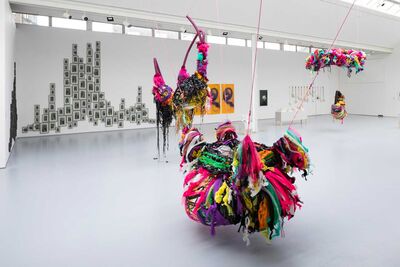 A gallery space in the foreground a colourful suspended sculpture in the background images visible on a wall