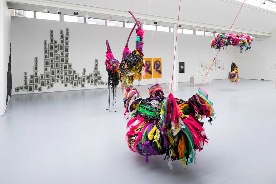 A gallery space in the foreground a colourful suspended sculpture in the background images visible on a wall
