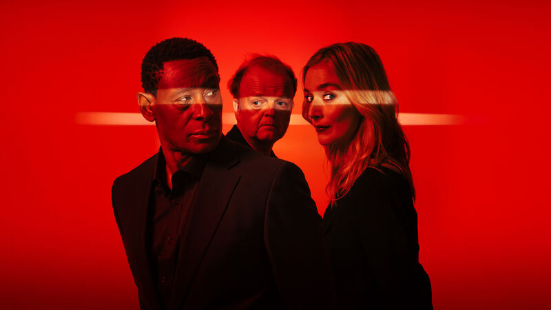 Three people in black suits stand unsmiling against a red backdrop.