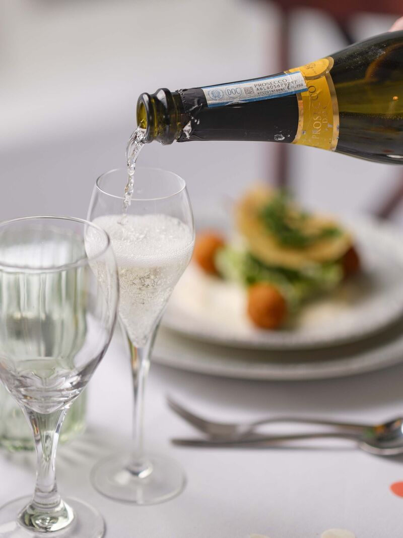 Prosecco being poured into a glass. Behind  a set table and full meal is slightly out of focus. 