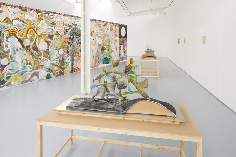 A hung textile work with watercolour painting and graphic text hangs to the right. In the foreground is a plinth with a textile piece. The colours are muted and soft, the gallery is naturally lit and bright. 