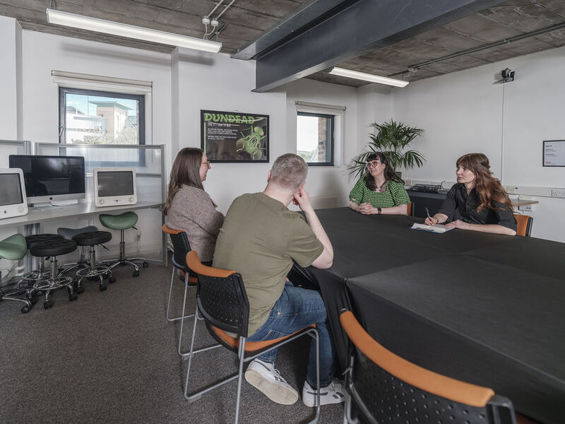 People sit in a meeting room. There are old style computer monitors on the desk in the left corner and a plant on the right