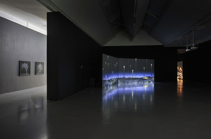 This wide shot photograph shows an installation in gallery two, with two photographs on a grey wall to the left of the image. The rest of the gallery is boxed in to make a dark space where projected films are shows. The film we can see shows a blue glowing landscape with bright lights in a dark sky. 