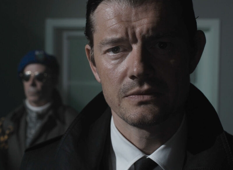 Still from Bulk of a mans face. Another man stands in the background wearing a hat and sunglasses