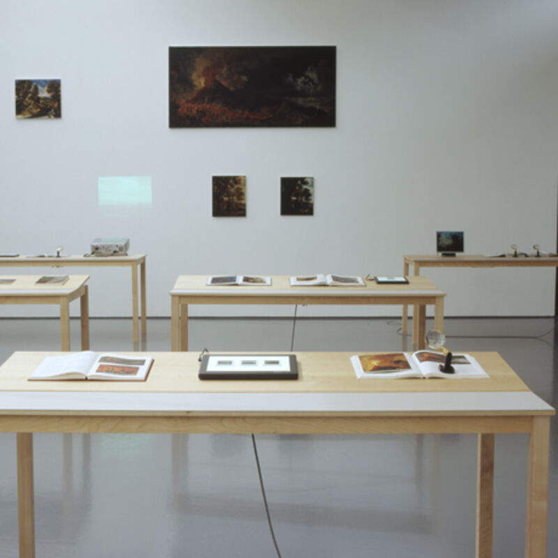 From Matts Leiderstam's exhibition in DCA Galleries. Four tables stand in the galleries with books and framed paintings on them. On the walls, there are four landscape paintings.