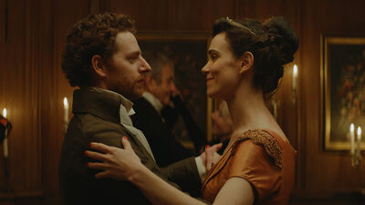 A man and woman in regency dress dance in a ballroom.