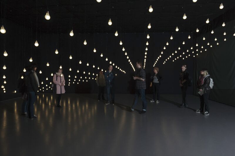 From Jim Campbell's exhibition. Visitors to DCA Galleries walk in a dark room, beneath tens of white light bulbs which are hanging from the ceiling.