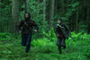 A man and a young boy run through a green forest holding crossbows.