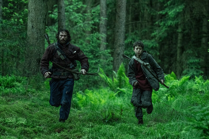 A man and a young boy run through a green forest holding crossbows.