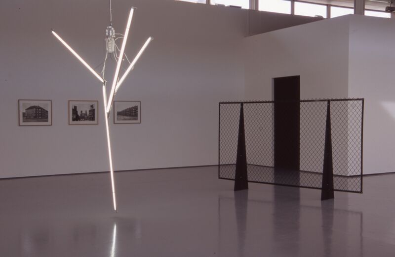 An abstract white sculpture made of strip lights sits in DCA Galleries, next to framed black-and-white photographs of buildings, and a fence made out of barbed wire.