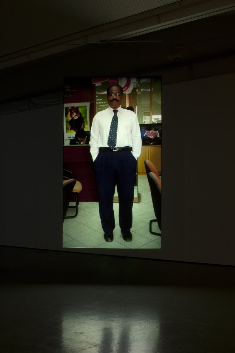 An image from Nikolaj Bendix Skyum Larsen's exhibition in DCA galleries. A man wearing a dress shirt, tie, and trousers stands in an office environment.