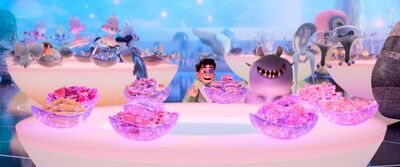 A little boy and a worm-like monster with a smiley face look at a table covered in sparkling food.