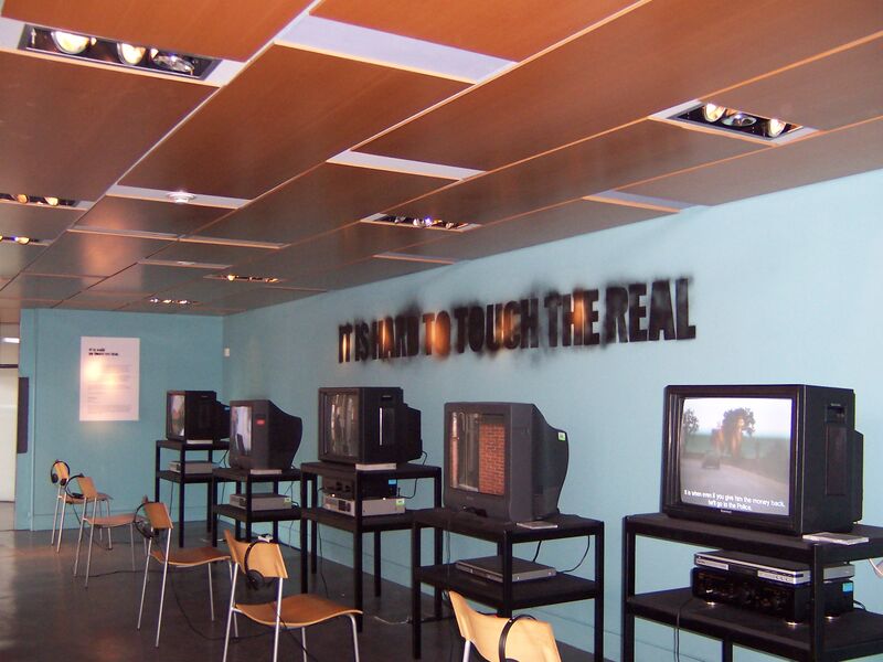 Chairs face televisions that each playing different videos. The walls are blue with the words 'It Is Hard to Touch the Real' spray painted on them.