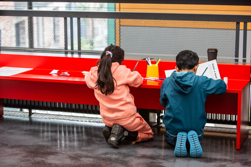 two children crouch down doing drawing leaning on a red bench a yellow pot of pens is infront of them 