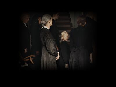 A solemn Victorian child in a black dress stands amongst women in black dresses